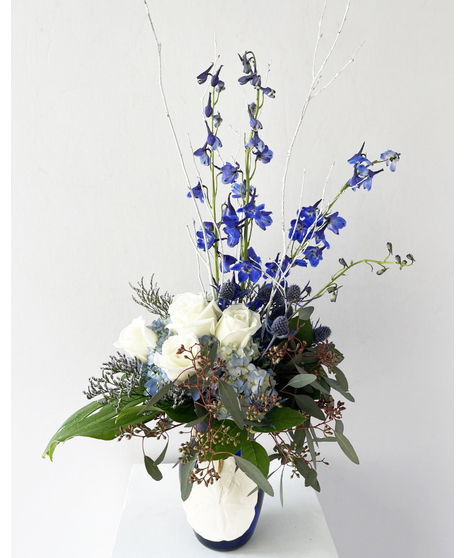 Blue delphinium, white hydrangea and white roses in a sapphire blue vase with sand dollar accent.
