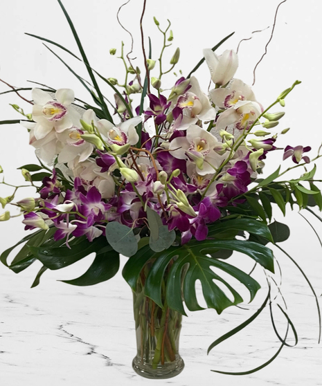 Glass vase of orchid in purple and white with curly willow and tropical greens.
