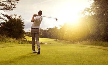 A golfer swings a club on a lush green course with trees and a glowing sun in the background.