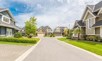 Houses line a wide, clean street in a suburban neighborhood under a partly cloudy sky, with neatly trimmed lawns and gardens.