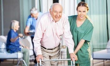 An elderly man using a walker is assisted by a smiling female caregiver in green scrubs. In the background, two other elderly individuals are seated and engaged in activities.