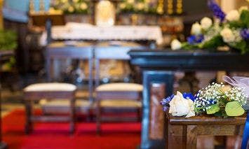 Flowers rest on a wooden pew in a decorated altar during a church ceremony, with two empty chairs in front and a red carpet below.