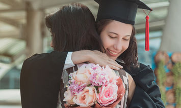 A graduate in a cap and gown embraces another person holding a bouquet of pink and white flowers, within an outdoor setting featuring a structure with beams.